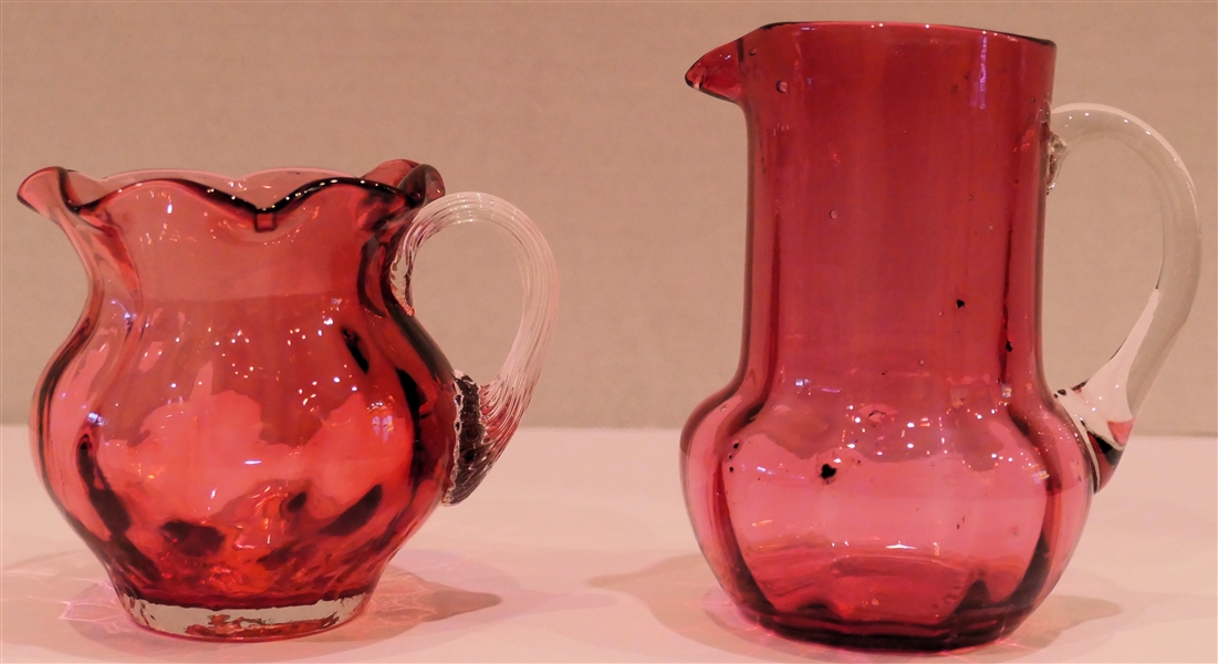 2 Hand Blown Pilgrim Glass Cranberry Pitchers  - Squatty Pitcher with Clear Ribbed Applied Handle Has Original Paper Label Other Pitcher with Smooth Clear Applied Handle Measures 4 1/2" Tall 